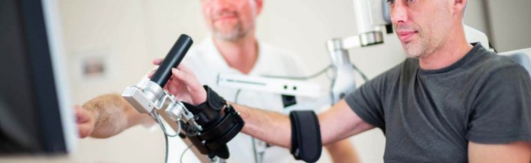 Robotikgestützte Armtherapie: Patient macht Übungen am PC, Therapeut sitzt daneben