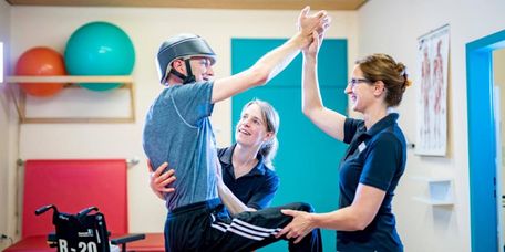 Patient mit Kopfverletzung und Helm bei der Therapie mit zwei Therapeutinnen