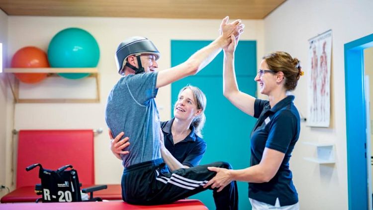 Patient mit Kopfverletzung und Helm bei der Therapie mit zwei Therapeutinnen