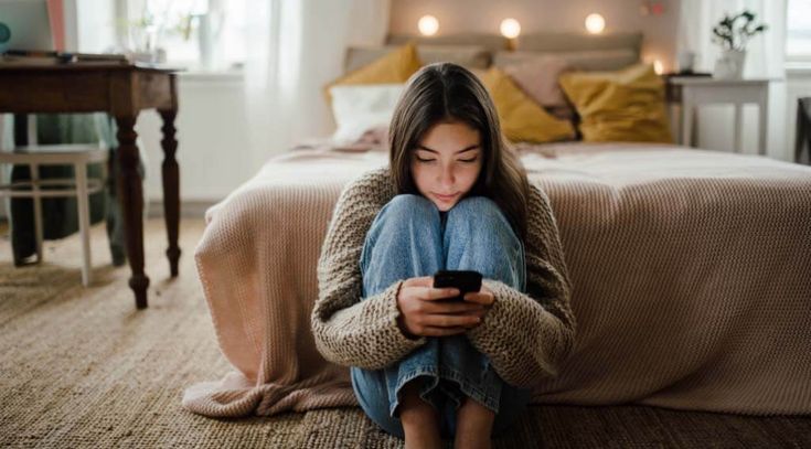 Mädchen sitzt auf dem Boden vor dem Bett und schaut in ihr Handy