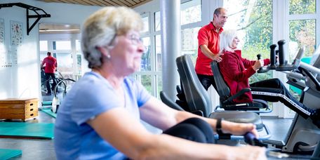 Ältere Dame am Rudergerät in der Reha, im Hintergrund sieht man einen Therapeuten mit einer Patientin am Gerät