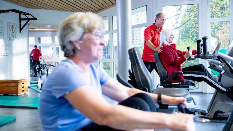 Ältere Dame am Rudergerät in der Reha, im Hintergrund sieht man einen Therapeuten mit einer Patientin am Gerät