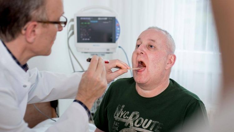 Arzt untersucht bei einem sitzenden Patienten den Mund- und Rachenraum im Rahmen einer Schluckuntersuchung