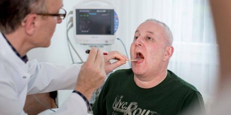 Arzt untersucht bei einem sitzenden Patienten den Mund- und Rachenraum im Rahmen einer Schluckuntersuchung