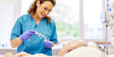 Pflegefachkraft in blauer Arbeitskleidung unterstützt einen älteren Patienten im Bett bei einer Maßnahme zur Schluckunterstützung im Mund- und Rachenbereich.