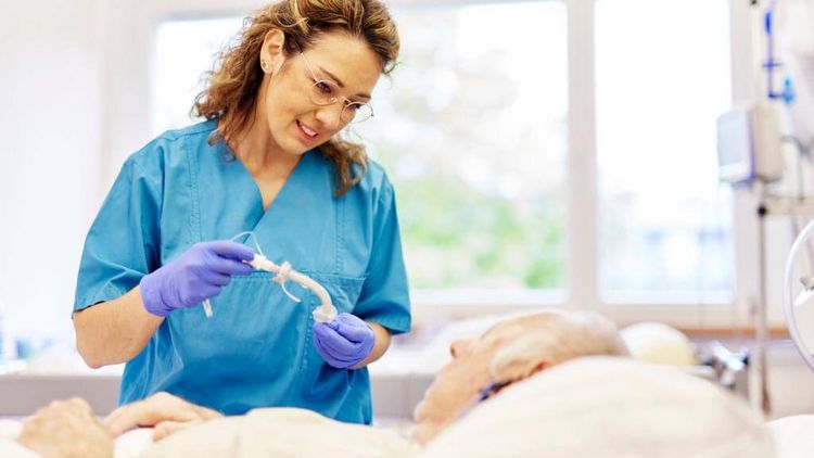 Pflegefachkraft in blauer Arbeitskleidung unterstützt einen älteren Patienten im Bett bei einer Maßnahme zur Schluckunterstützung im Mund- und Rachenbereich.