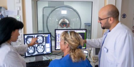 Ärzte begutachten MRT-Bilder vom Gehirn am Bildschirm, im Hintergrund liegt ein Patient im MRT-Gerät