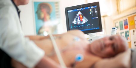 Arzt führt eine Echokardiografie bei einem Patienten aus, im Hintergrund sieht man den Bildschirm