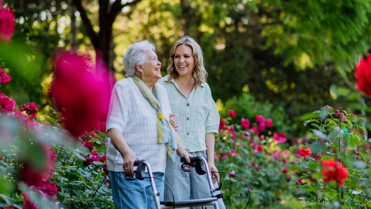 Eine Oma geht mit ihrer erwachsenen Enkelin in einem Garten mit blühenden Blumen spazieren