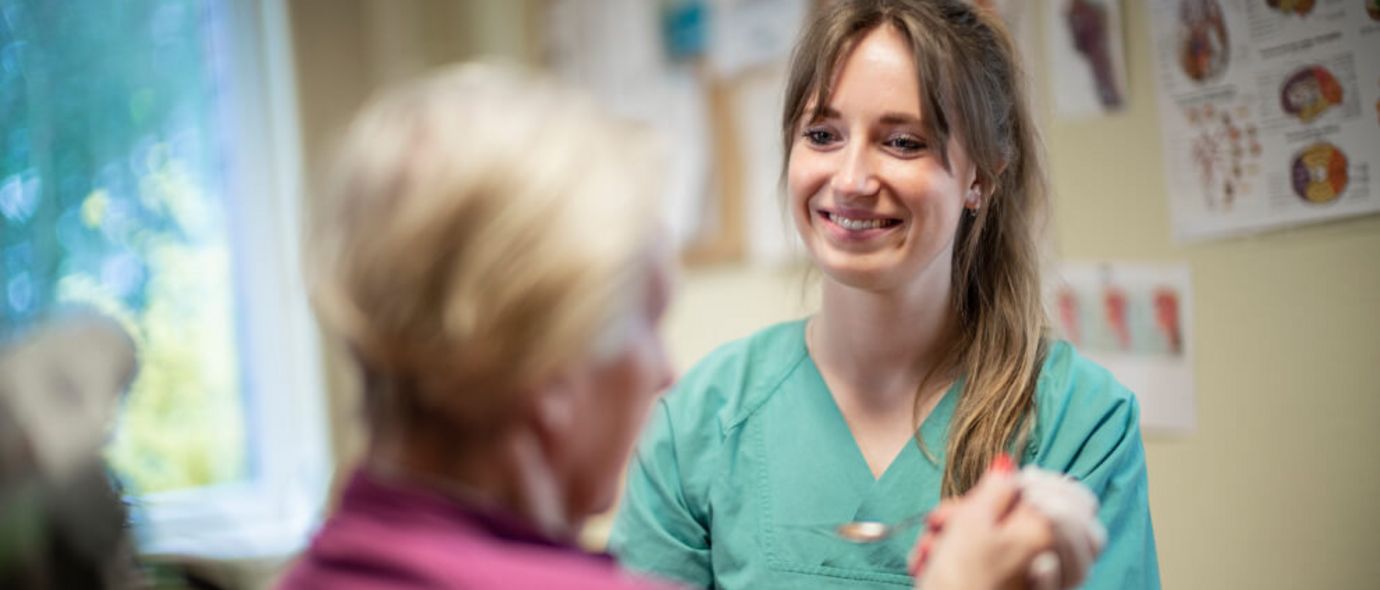 Eine Pflegerin reicht einer Patientin im Rollstuhl eine Löffel