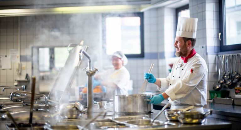 Ein Koch rührt in einer Großküche mit einem Schneebesen in einem dampfenden Topf