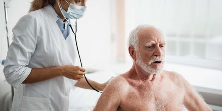 Ärztin in weißem Kittel untersucht älteren Patienten mit einem Stethoskop am Rücken