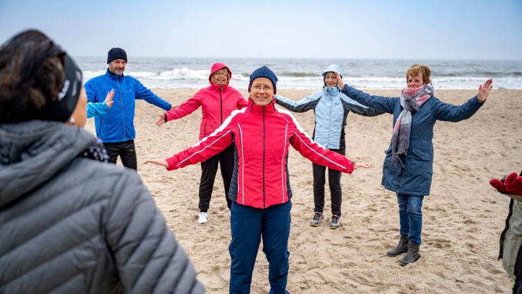 Therapeutin und Patientengruppe wärmen sich im Herbst am Strand mit Gymnastik auf