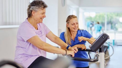Patientin sitzt auf einem Ergometer, Therapeutin steht daneben und zeigt auf die Anzeige