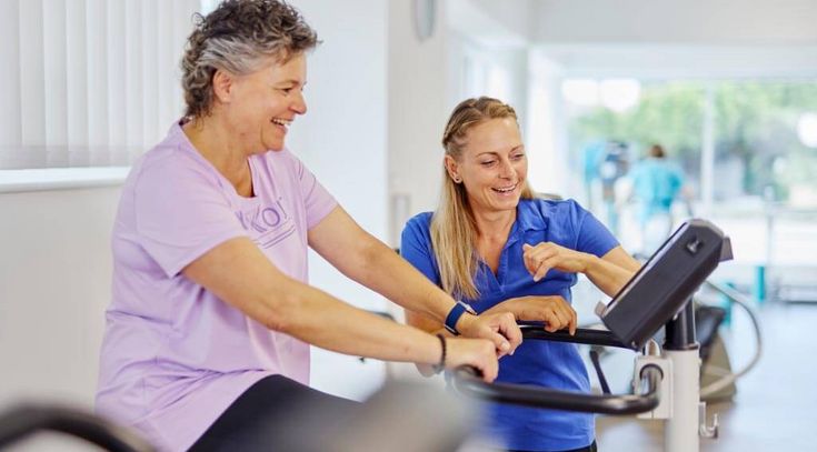 Patientin sitzt auf einem Ergometer, Therapeutin steht daneben und zeigt auf die Anzeige