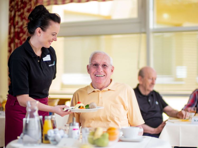 Eine Servicekraft serviert einem älteren fröhlichen Patienten Essen am Tisch