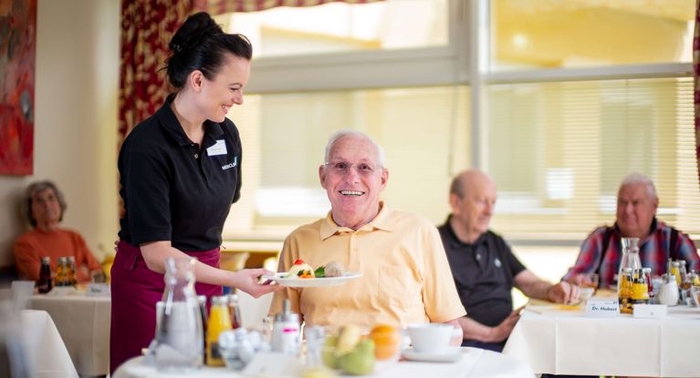 Eine Servicekraft serviert einem älteren fröhlichen Patienten Essen am Tisch