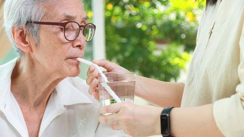 Eine ältere Frau trinkt mithilfe eines Trinkhalms Wasser, während eine Betreuungsperson das Glas zur Schluckunterstützung hält.