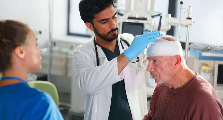 Arzt untersucht älteren Patienten mit Kopfverband, weibliche Pflegekraft steht daneben