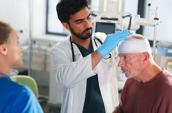 Arzt untersucht älteren Patienten mit Kopfverband, weibliche Pflegekraft steht daneben