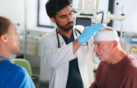 Arzt untersucht älteren Patienten mit Kopfverband, weibliche Pflegekraft steht daneben