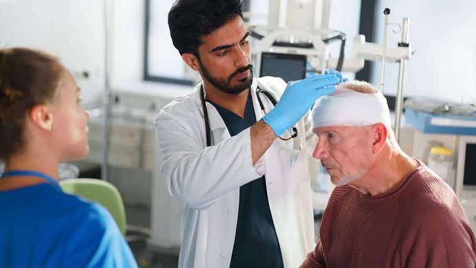 Arzt untersucht älteren Patienten mit Kopfverband, weibliche Pflegekraft steht daneben