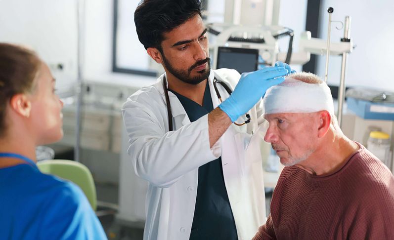 Arzt untersucht älteren Patienten mit Kopfverband, weibliche Pflegekraft steht daneben