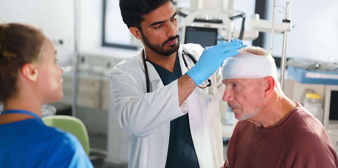 Arzt untersucht älteren Patienten mit Kopfverband, weibliche Pflegekraft steht daneben