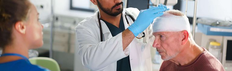 Arzt untersucht älteren Patienten mit Kopfverband, weibliche Pflegekraft steht daneben