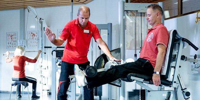 Männliche Person beim Krafttraining in der Reha, Therapeut unterstützt
