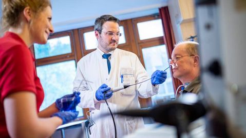 Arzt und Therapeutin führen bei einem sitzenden älteren Patienten eine endoskopische Untersuchung zur Diagnostik von Schluckstörungen durch.