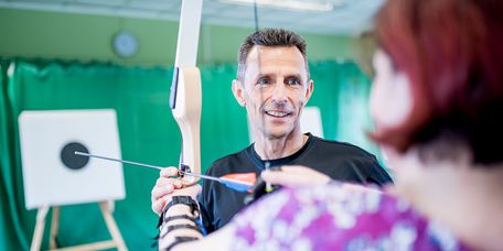 Therapeut unterstützt Patientin in der Sporthalle beim Bogenschießen