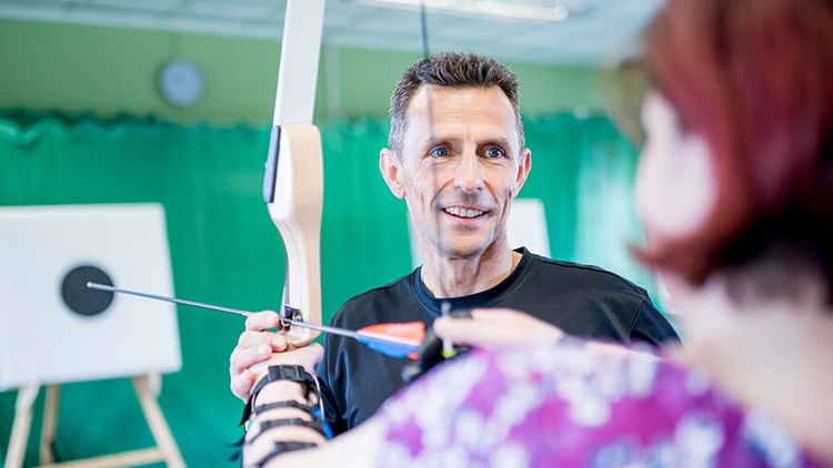 Therapeut unterstützt Patientin in der Sporthalle beim Bogenschießen