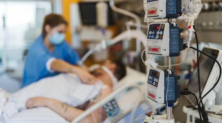 Patient liegt im Krankenhausbett auf der Intensivstation, Pflegekraft kümmert sich um ihn, im Vordergrund sind Geräte und Schläuche zu sehen