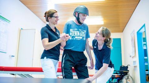 Patient mit Kopfverletzung in der Reha, er trägt einen Helm, zwei Therapeutinnen helfen im beim Gehen