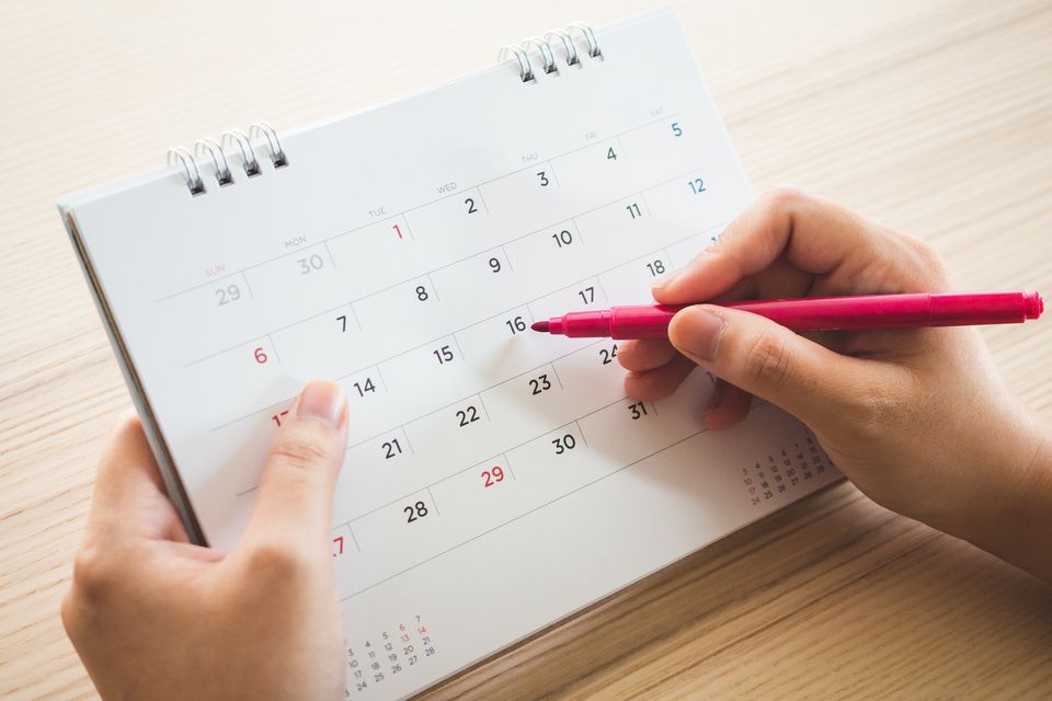 Eine Person markiert mit einem pinkfarbenen Stift ein Datum in einem Tischkalender. Der Kalender zeigt einen Monatsüberblick mit Zahlen von 1 bis 31, wobei die Sonntage in rot markiert sind. Die Person hält den Kalender mit einer Hand fest, während sie mit der anderen Hand den Stift führt.